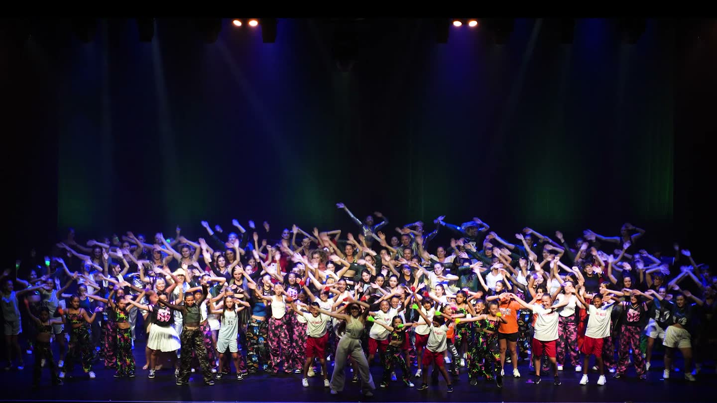 Tous les groupes du Studio M DANCE réunis sur scène au salut, bras levés, sous les projecteurs.