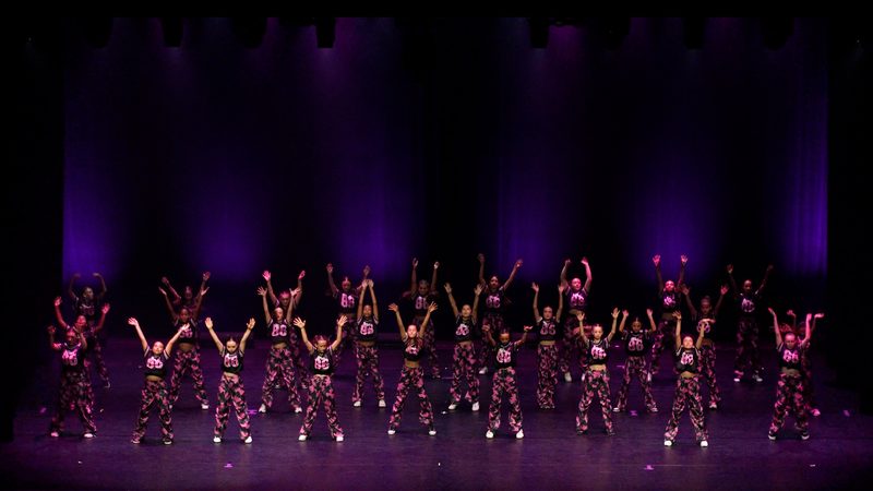 Un grand groupe d'enfants en costumes à paillettes, bras levés, sous une lumière violette.