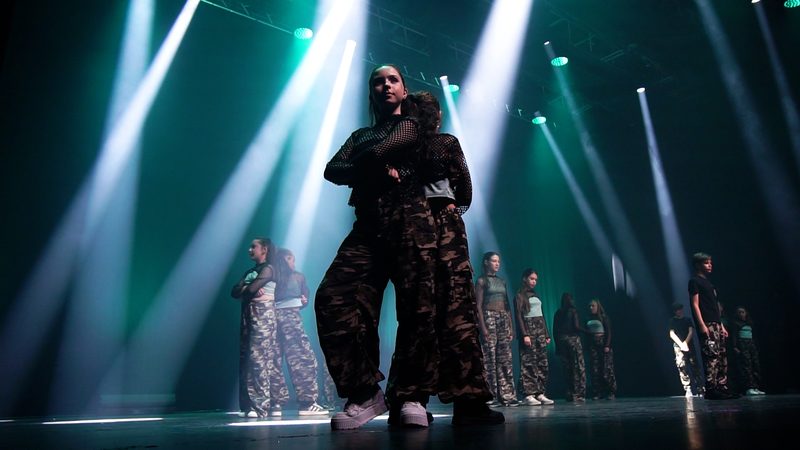 Une danseuse au premier plan en tenue camouflage, devant son groupe, sous des faisceaux blancs.
