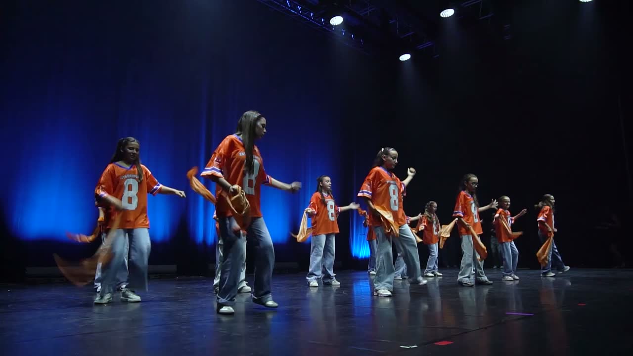 Le groupe hip-hop des 11-12 ans en maillots orange et jeans clairs, devant un fond bleu.